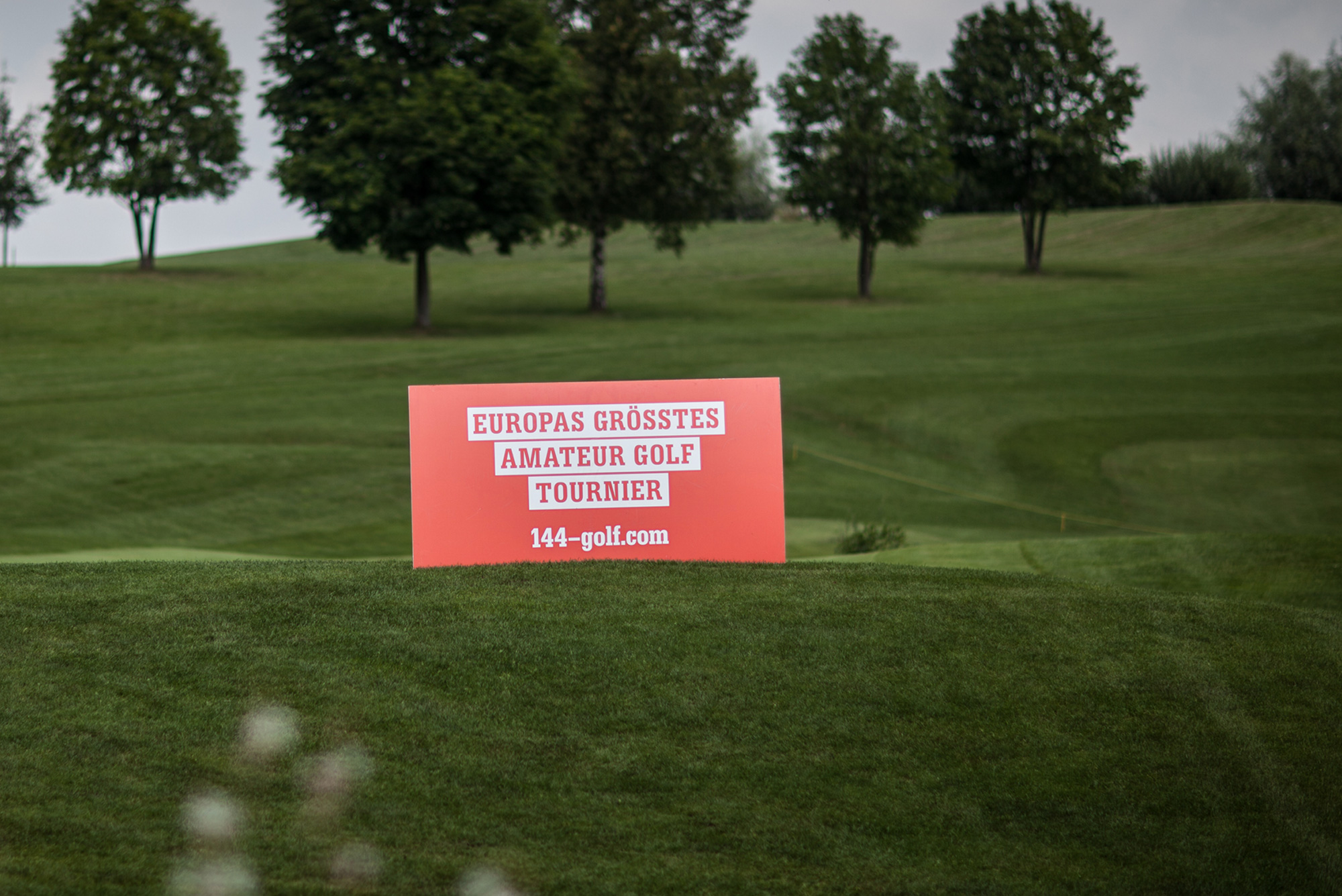 Foto einer grünen Golfrasenlandschaft mit Bäumen die im Vordergrund die lachfarbene Karte des Amateur Golf Turniers zeigt. 